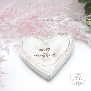 A white, heart-shaped decorative object inscribed with "Happy everything" is placed against a soft, light pink and green floral background. This charming piece adds a touch of happiness to any space, perfect for enhancing your home decor. The Mango Wooden Heart Trays with Sentiments are ideal for this purpose.