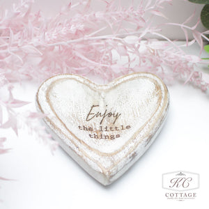 A rustic Mango Wooden Heart Trays with Sentiments piece with the words "Enjoy the little things" inscribed on it. The heart is set against a soft background with delicate pink foliage in the corner.
