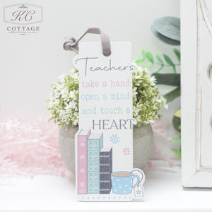 A decorative bookmark with the text: "Teachers take a hand, open a mind, and touch a heart." It features illustrations of three stacked books, a blue polka-dotted mug, and a small plant. The background boasts soft pastel colors, making it an elegant piece for your home's decor. Introducing the Teacher Gift - Thank You Bookmark.