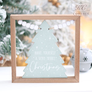 The Christmas Tree Sign with Beads features a wooden frame surrounding a light green tree design. White text on the tree reads "Have Yourself a Very Merry Christmas," and it is enhanced with bead tinsel, set against a blurred, snowy background accented by fir branches.