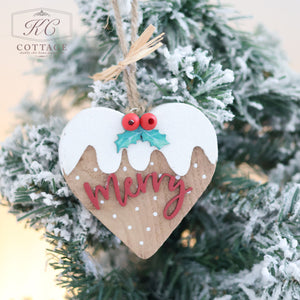 The Christmas Pudding Hanging Hearts adorn a snowy Christmas tree, featuring a heart-shaped wooden design with white icing, red berries, and the word "Merry" in red. Resembling a Christmas pudding and suspended by a twine loop, this ornament radiates cozy and festive charm.