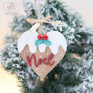 A Christmas Pudding Hanging Heart, featuring a wooden heart-shaped design with white frosting-like details and "Noel" written in red, is displayed on a snowy pine branch. Red berries and green leaves attached with twine adorn the top, adding to its festive charm.