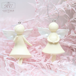 Two cream-colored Large Hanging Angels with simple, faceless designs and white wings hang by silver strings. They are surrounded by light pink, fluffy foliage, creating a soft, ethereal backdrop that enhances your home decor. The logo "KC Cottage" is in the top left corner.