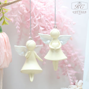 Two cream-colored Large Hanging Angels with white wings hang from silver cords. The one on the left holds a heart close to its chest, while the one on the right carries a small bowl. Perfect for home decor, both are set against a soft, pink, floral backdrop.