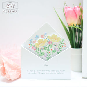 A delicate greeting card for a grandmother showcases pastel-colored flowers inside a Wooden Floral Envelope Flower Boxes with the name "Nan" written on it. Nearby, amidst charming home decor, there is a pink tulip. A heartfelt message reads, "If I had a flower for every time you made me smile, I'd have a garden to walk in.