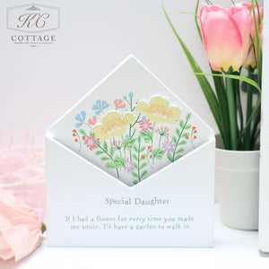 A wooden floral envelope flower box labeled "Special Daughter" is adorned with colorful, illustrated flowers and nestled in an envelope. An orange tulip in the background adds a touch of nature. Perfect even as home decor, the box bears the message: “If I had a flower for every time you made me smile, I’d have a garden to walk in.”