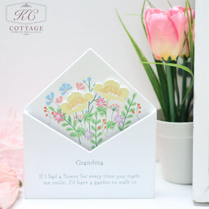 A decorative card with colorful flowers displayed in a wooden floral envelope flower box. The card holder is inscribed with "Grandma - If I had a flower for every time you made me smile, I'd have a garden to walk in." Beside it, enhancing your home decor, is a pink tulip in a white pot.