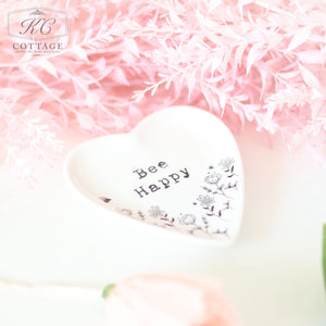 A heart-shaped dish with the words "Bee Happy" and floral illustrations sits on a white surface. Surrounding the dish are pink, feathery artificial flowers, adding charm to your home decor. A single pink rose is also visible in the bottom left corner of the Gift Set - Thinking Of You.