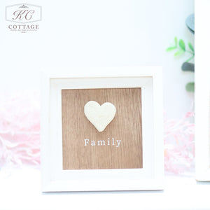 A small framed sign with a white frame and a wooden background. The wooden background features a white heart above the word "Family" in white text. This charming piece of home decor, called the Pebble Heart Sign, is part of a setup with blurred greenery and a decorative pink ribbon in the background.