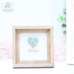 A small framed art piece with a beige wooden frame. Inside, a greenish-blue heart made of textured material is centered above the word "Peace" in simple black font. The background is a light, subtle shade, perfect for home decor. The logo "KC Cottage" is at the top left corner of the Pebble Heart Sign.