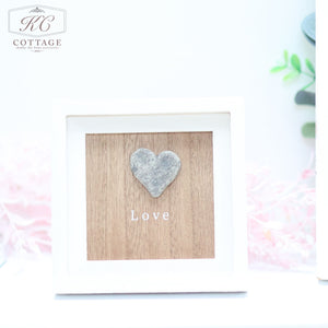 A framed decoration from KC Cottage featuring a wooden background with a raised heart shape and the word "Love" written in white beneath it. This charming Pebble Heart Sign has a white frame with soft pink flowers and green foliage softly blurred in the background.