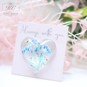A Glass Spring Floral Heart with Sentiment Card, featuring a heart-shaped, clear resin accessory with blue flowers inside. The card is placed against a softly blurred, pink floral background—perfect for any home decor setting.