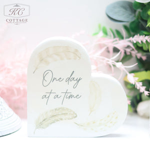 A Grey Feather Ceramic Memory Heart Block with the text "One day at a time" written on it. It is adorned with light-colored feather illustrations and is set against a background featuring soft pink and green foliage, perfect for adding a touch of elegance to your home decor.