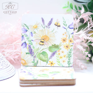 A Daisy Bee Floral Coaster featuring a watercolor design of various flowers and two bees is propped up against a stack of similar coasters. Perfect for home decor, the flowers include daisies, lavender, and others in shades of yellow, purple, and green. The background has pink-frosted foliage.