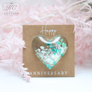 A small square card doubles as charming home decor, featuring a heart-shaped dome filled with delicate white flowers and greenery. The Wedding Pocket Heart Token On Card reads "Happy ANNIVERSARY" in elegant text. Soft, pastel pink flowers surround the card, creating a romantic and gentle atmosphere.