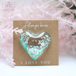 A heart-shaped glass ornament featuring white floral designs rests on a brown card. The card reads "Always know I LOVE YOU" in white text. Pink foliage surrounds the card, adding a soft, romantic touch. This Wedding Pocket Heart Token On Card makes for exquisite home decor.