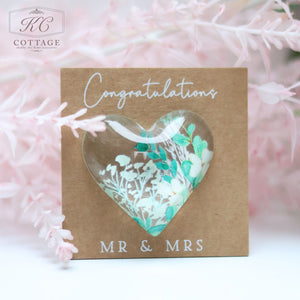 A **Wedding Pocket Heart Token On Card** with "Congratulations MR & MRS" written on it. The card features a heart-shaped window displaying a floral design with white and blue flowers. The background has delicate pink foliage, perfect for incorporating into your home decor.