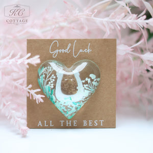 A small, heart-shaped cutout card with "Good Luck" written at the top and "All the Best" at the bottom, featuring pastel floral designs and delicate pink branches in the background. The Wedding Pocket Heart Token On Card has a transparent heart window with a glimmering interior, perfect for home decor.