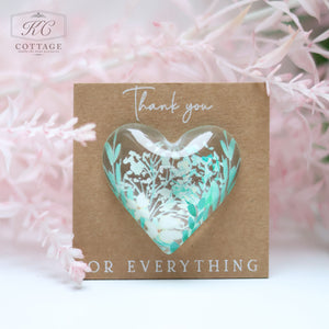 A **Wedding Pocket Heart Token On Card** with a transparent heart-shaped window containing a floral design, featuring green and white leaves and flowers. The brown card, perfect for home decor, has the text "Thank you" at the top and "FOR EVERYTHING" at the bottom. The background includes pink foliage.