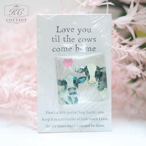 A small card features an illustration of two cows with a pink heart above them. The text above reads, "Love you til the cows come home," and the text below says, "Here's a little pocket hug just for you. Keep it as a reminder of how much I care, for the times when I cannot be here." This charming Pocket Hugs with Sentiments also makes a lovely piece of home decor.
