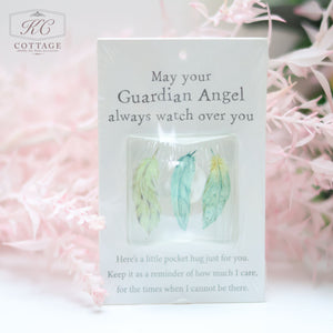 A charming Pocket Hugs with Sentiments with three pastel feather charms. Text reads: "May your Guardian Angel always watch over you. Here’s a little pocket hug just for you. Keep it as a reminder of how much I care, for the times when I cannot be there." Background features pink foliage, perfect for home decor.