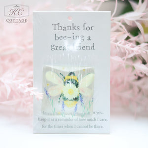 A Pocket Hugs with Sentiments with text "Thanks for bee-ing a great friend" and a small plush bee attached. The card, surrounded by pink, out-of-focus flowers, reads under the bee: "Here’s a little pocket hug just for you. Keep it as a reminder of how much I care, for the times when I cannot be there." Perfect for home decor.