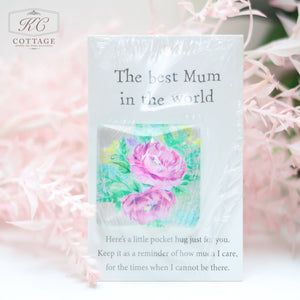 A card with the phrase "The best Mum in the world" printed on it. Below, a small fabric patch with pink roses is attached. The bottom text reads, "Here's a little pocket hug just for you. Keep it as a reminder of how much I care, for the times when I cannot be there." Perfect for home decor, Pocket Hugs with Sentiments has a soft background with pink foliage and is wrapped