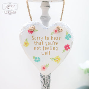 A Bright Floral Slate Hanging Hearts home decor piece with a white background and floral accents. The text on the heart reads, "Sorry to hear that you're not feeling well." It is hanging by a twine string against a light-colored background.