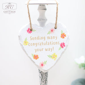 A heart-shaped decorative plaque with a jute string reads "Sending many congratulations your way!" in gold letters. Featuring colorful floral designs around the text, this charming piece of home decor hangs from a white, ornate stand.