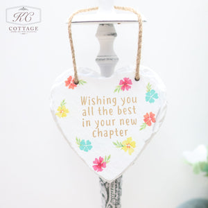 A Bright Floral Slate Hanging Hearts with a rope for hanging features colorful flowers and the message: "Wishing you all the best in your new chapter." The piece of home decor hangs from a white hook, with the background softly out of focus.