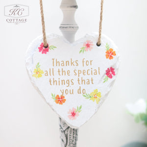 A heart-shaped home decor piece adorned with floral designs and the text "Thanks for all the special things that you do" written on it. The Bright Floral Slate Hanging Hearts decoration hangs from a twine on an ornate stand with a blurred background.
