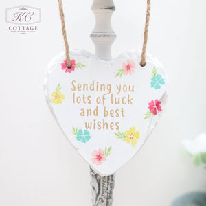 A heart-shaped ornament made of a white material hangs from a brown string. The home decor piece is adorned with colorful flowers and bears the message: "Sending you lots of luck and best wishes." The background is white with a partial view of a light fixture. This charming item is called Bright Floral Slate Hanging Hearts.