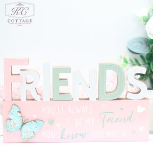 A charming piece of home decor, the Bright Butterfly Wooden Signs - Family, Friend, Home features pastel-colored letters spelling "FRIENDS" and a pink base with the touching phrase "You'll always be my friend you know too much." Adorned with a butterfly and hearts.