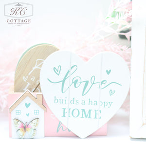 A decorative white heart-shaped sign reads "Love builds a happy home" in green cursive. It is placed next to a Bright Butterfly Wooden Coasters Set with a butterfly design and pink roof. The background features soft pink foliage, adding a charming touch to this delightful piece of home decor.