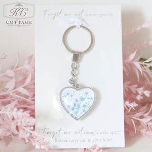 A Glass Forget Me Not Heart Keyring is displayed against a white card with text. The card reads, "Forget me not as time goes by" and "Forget me not though we’re apart, forever carry me in your heart." Pink flowers are in the background, complementing home decor accents.