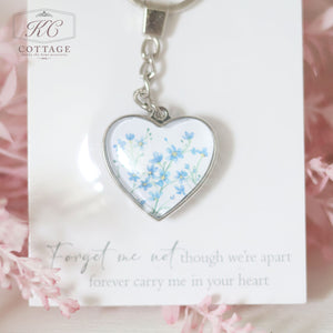 A Glass Forget Me Not Heart Keyring featuring delicate blue forget-me-not flowers is attached to a card with the message: "Forget me not, though we're apart, forever carry me in your heart." Perfect as a piece of home decor, the card and keyring are set against a soft pink, floral background.