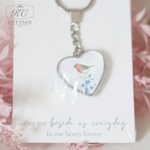 A Glass Forget Me Not Heart Keyring with a small red and brown bird and blue flowers. The keychain is placed on a card that reads, "...they stay beside us every day in our hearts forever." The background features soft pink crinkled paper, making it a charming addition to your home decor.