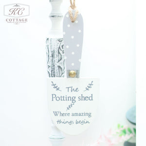 A small decorative Wooden Garden Trowel Hanger, perfect for home decor, hangs by a rope. The blade is white with "The Potting Shed - Where amazing things begin" written in gray. The handle is painted gray with white polka dots. In the background, a decorative candle holder and soft focus greenery add charm.