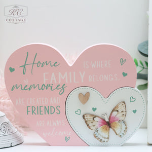 A decorative pink heart-shaped sign with a smaller heart attached, perfect for home decor. The large heart reads, "Home is where family belongs, memories are created and friends are always welcome." The smaller heart features a wooden butterfly with colorful wings and heart motifs around it. Presenting: Bright Butterfly Heart Plaques.
