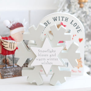 The Christmas Snowflake with Pastel Star ornament, featuring the heartwarming message "Snowflake kisses and warm winter wishes," is displayed against a blurred background adorned with festive holiday elements, including a bird in a Santa hat and a sign reading "Made with Love," spreading cheer all around.