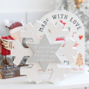 A close-up of a decorative item, the Christmas Snowflake with Pastel Star, featuring the inscription, "If snowflakes were kisses, I'd send you a blizzard." In the background, festive decorations include a sign that says "MADE WITH LOVE" and a blurred view of a Santa figurine, enhancing the holiday atmosphere.
