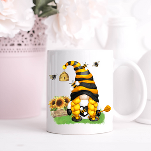 A Bee & Sunflower Gonk Mug features a whimsical illustration of a gnome with yellow braids and a black-and-yellow striped hat, standing on grass. The gnome is surrounded by bees, a sunflower, a beehive, and an overturned honey pot. Perfect for adding charm to your home decor.