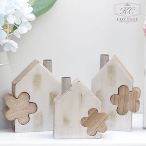 The image shows three small, white, house-shaped wooden decorations with wooden clover cutouts attached to their fronts. They are placed on a white surface with part of a white vase and a bouquet of light-colored flowers visible in the background. Perfect for enhancing your home decor, this Wooden House Set with Flower Block adds a charming touch.