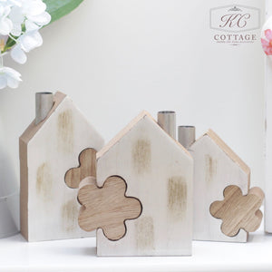 A decorative Wooden House Set with Flower Block features three wooden house-shaped pieces with cut-out designs. One house has a heart cut-out, the second has a flower, and the third has an abstract shape. They are light-colored with a rustic finish, set against a white background.