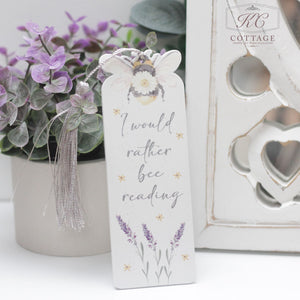 A white bookmark with a tassel displays the text "I would rather bee reading" accompanied by a bee illustration and lavender flowers. Set against a backdrop of purple blooms and decorative heart-shaped cutouts, this charming Daisy Bee Bookmarks piece doubles as delightful home decor.