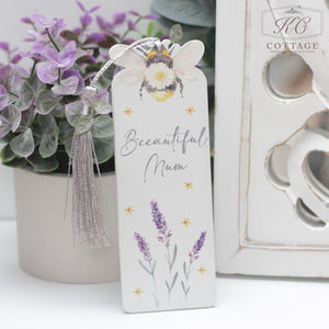 A decorative Daisy Bee Bookmarks with "Beeautiful Mum" written on it, adorned with illustrations of a bee, yellow flowers, and lavender flowers. It has a grey tassel attached to the top. Perfect for adding charm to your home decor, the bookmark is placed next to a small purple flower arrangement and a decorative white object.