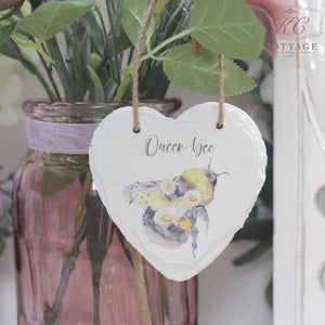 A Bee Slate Hanging Hearts with an illustration of a bee and the text "Queen Bee" hangs by a string in front of a glass jar containing a bouquet with purple and white flowers. The jar, tied with a purple ribbon, adds charm to your home decor. The background features leaves and part of a white structure.