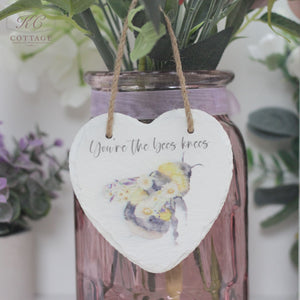 A Bee Slate Hanging Heart with an illustration of a bee and flowers hangs from a twine string. The text on the heart reads "You're the bees knees." This charming piece of home decor is attached to a pink glass vase containing various flowers and greenery.