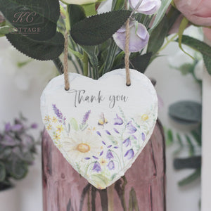 A Bee Slate Hanging Hearts made of white slate, adorned with colorful floral illustrations and the words "Thank you" written in the center, hangs from twine in front of a pink vase filled with purple and white flowers. This charming piece of home decor is set against a background of leaves and blurred flowers.