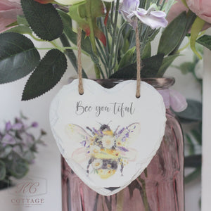 A decorative heart-shaped slate hangs by twine on a bouquet of flowers in a pink glass vase. The Bee Slate Hanging Hearts features an illustrated bee adorned with flowers and the text "Bee you tiful." The background includes leaves and flowers, adding a charming, rustic feel to this delightful piece of home decor.